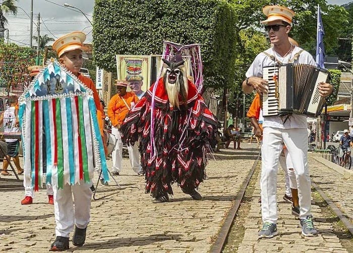 Mascarado de traje preto e vermelho ao lado de sanfoneiro, desfilando em via com trilho antigo