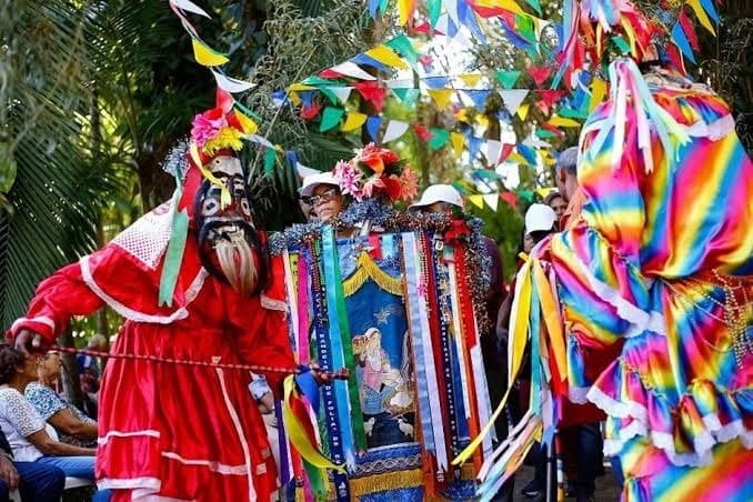 Mascarado da Folia de Reis ao lado da bandeira bordada com a imagem do santo, em meio a bandeirolas coloridas